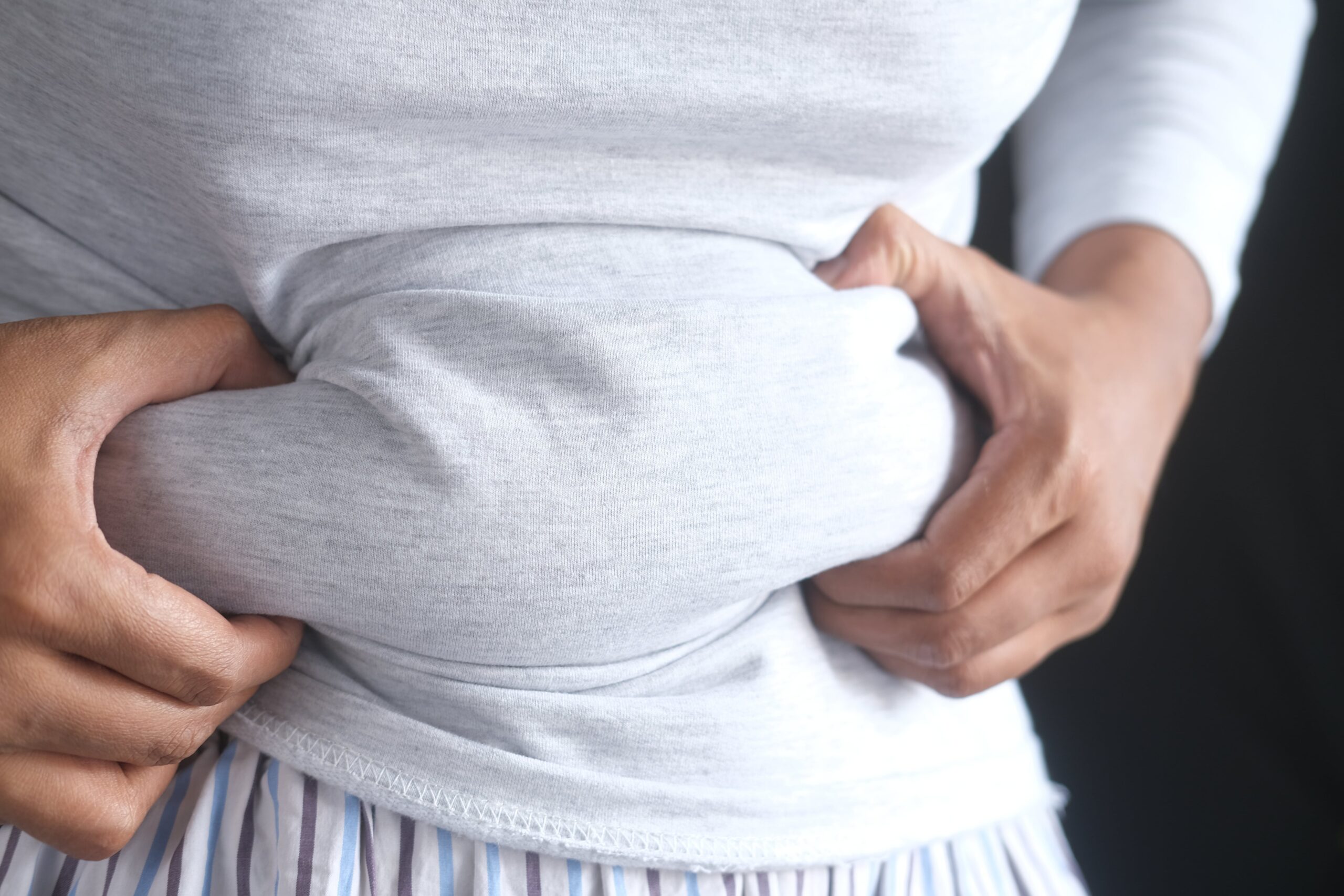 The image focuses on a close-up view of a person’s torso, specifically their abdomen, conveying a sense of discomfort or concern. The individual is wearing a light grey, long-sleeved t-shirt that is stretched taut across their belly, highlighting a noticeable fullness or potential weight gain. Their hands are clasped around their stomach, with fingers visibly digging into the soft tissue, suggesting they are either feeling pain, bloating, or self-consciously aware of their midsection.