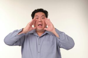 Man in a light blue shirt cupping his hands around his mouth as if shouting toward the camera against a neutral background.